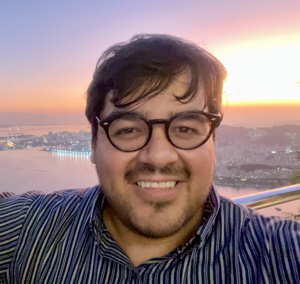 A smiling person wearing glasses with a bay and cityscape behind him