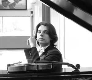 A person wearing a bow tie sits at a grand piano with its lid raised, and a violin resting on the piano.