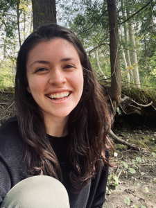 A smiling person with long hair poses in a wooded area