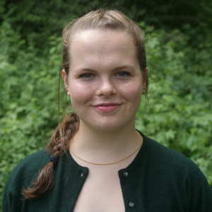 A smiling person stands in front of a green bush