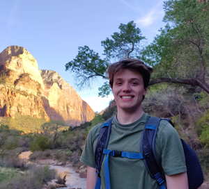 A smiling person wearing a backpack stands in front of a mountain and trees