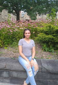 A smiling person sits on a stone wall with flowers, tree, and building behind her