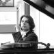 A person wearing a bow tie sits at a grand piano with its lid raised, and a violin resting on the piano.